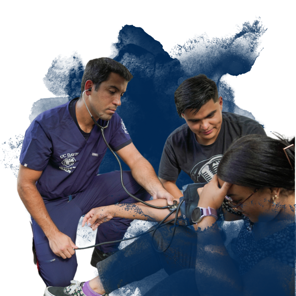 A healthcare professional in blue scrubs takes a person's blood pressure, with another individual observing.
