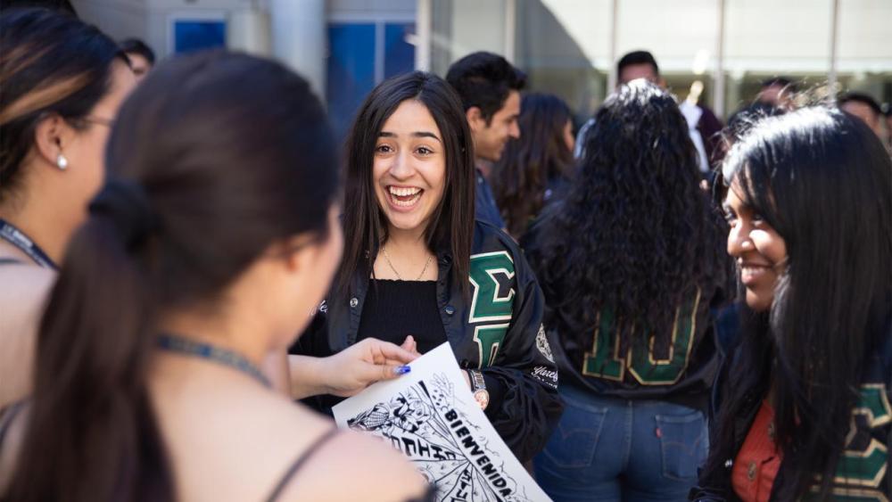 Female students in a group smiling with one of them holding a drawing that says "Bienvenida"