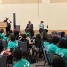 Chancellor May sits on stage in front of crowd of middle school and high school students seated at round tables wearing bright green tshirts. Ebony Lewis is at podium talking to crowd. 
