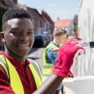 African American teenager wearing a red polo shirt, bright yellow construction vest and red gloves is painting a mural.