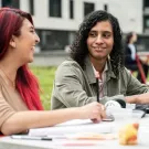 Woman with long hair dyed bright red sits at a table, laughing and engaged in conversation with man with shoulder-length curly hair who is working on a laptop. 