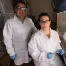 Fifth-year student Jasmine Diaz (seated right) and Professor Luis Carvajal-Carmona (standing left) pictured in lab wearing white lab coats, gloves and protective goggles.