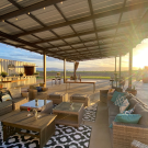 Couches and tables under a pergola overlooking a vineyard at sunset.