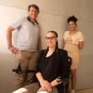 Man in stripped polo and khaki pants and woman in tan dress stand behind woman who is wearing a black top, glasses and isseated in a wheel chair. 