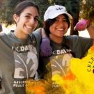 Two women wearing UC Davis Arboretum t-shirts each raise one arm to show strength. Their other arm is linked around the other person's waist. in corner of image is spiral of colors with Celebrating 35 years over a golden watercolor graphic.