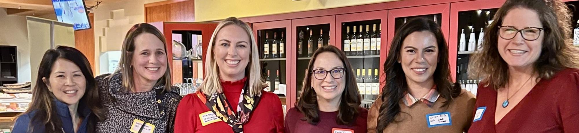 Six women stand together, smiling at the camera. They're in what appears to be a cafe or wine bar, with shelves of wine bottles visible behind them. The women are wearing a variety of colors, with the woman in the center wearing a bright red top. Most are wearing name tags.