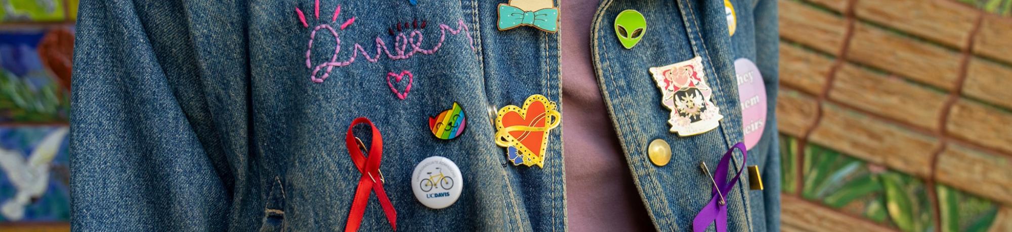 A person cropped from neck to waist wearing a jean jacket with stitching that reads "Queer," buttons that represent UC Davis, a red ribbon, a purple ribbon, a heart, a cat, a daisy, and a button that says "they, them, theirs"  